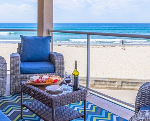 Oceanfront balcony with blue-cushioned wicker seating, wine, and snacks overlooking a sandy beach and surf.