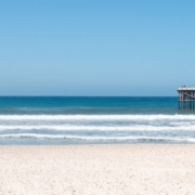 Sandy beach with rolling ocean waves and a wooden pier beneath a clear blue sky.