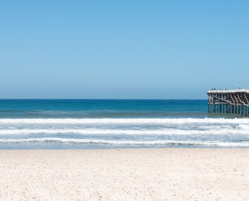 Sandy beach with rolling ocean waves and a wooden pier beneath a clear blue sky.